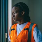 African American woman wearing PPE reflective vest, looking contemplatively out of window.
