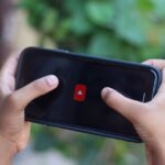 Close-up of hands holding a smartphone displaying a video app icon outdoors.