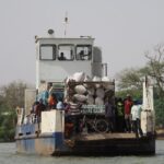 A ferry with passengers and goods travels across a river in Gambia, surrounded by lush greenery.
