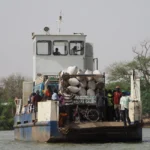 A ferry with passengers and goods travels across a river in Gambia, surrounded by lush greenery.