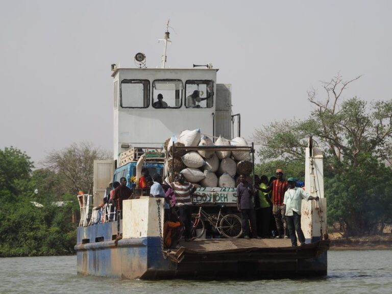 A ferry with passengers and goods travels across a river in Gambia, surrounded by lush greenery.