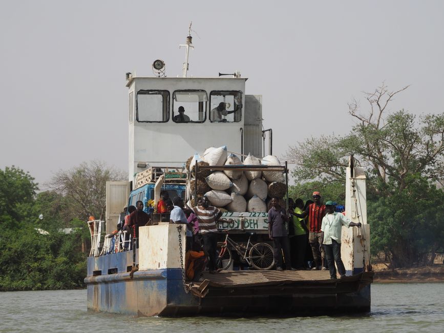 A ferry with passengers and goods travels across a river in Gambia, surrounded by lush greenery.